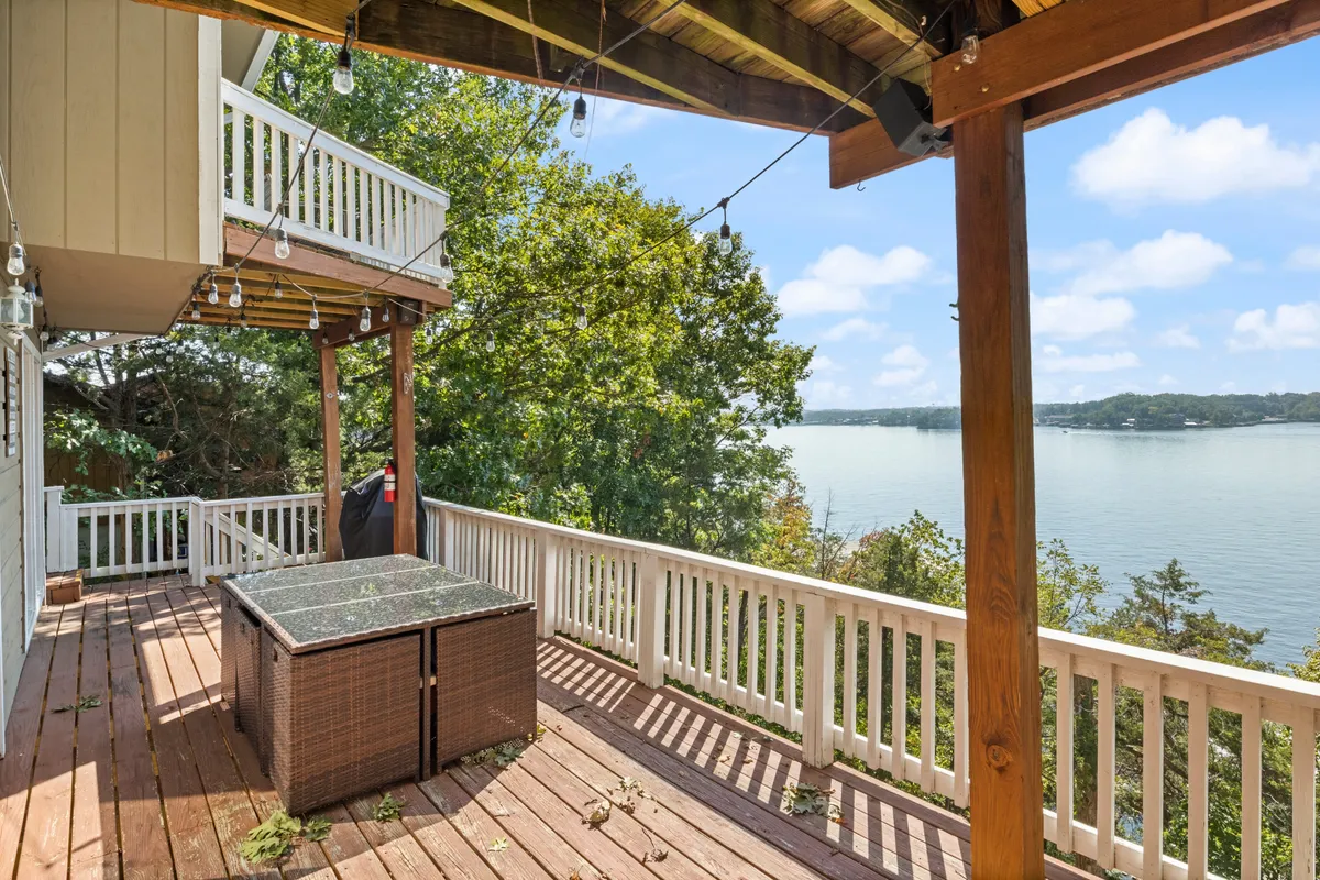 The image shows a spacious deck with a hot tub, ambient hanging lights, and a panoramic lake view stretching out beyond the railing. Outdoor dining furniture is set for gatherings, and the lighting suggests a relaxing, inviting atmosphere perfect for sunset cocktails or evening entertaining. The lake and sky are visible in the background, reinforcing the connection to nature and the scenic vistas that the property offers.