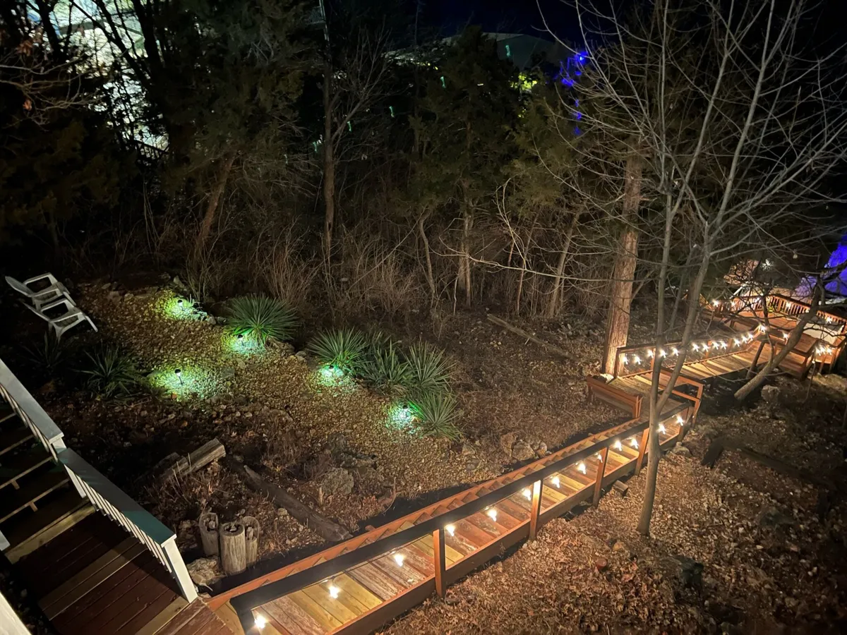 A serene nighttime view of a wooden pathway illuminated by garden lights, leading to a cozy seating area outdoors. The lighting suggests attention to safety and ambiance, while the setting feels inviting for evening strolls or gatherings.