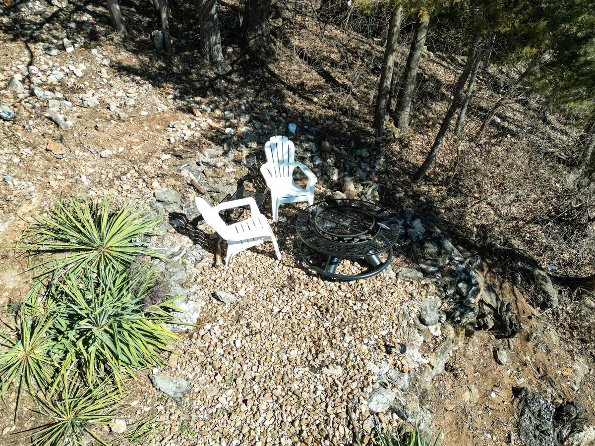 This cozy outdoor firepit area features two white chairs set on rocky ground amidst lush greenery, perfect for gathering around the fire and enjoying the lakeside ambiance under the stars.