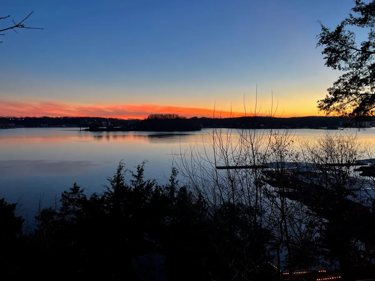 A panoramic view of the tranquil lake, featuring an island, sunset colors reflected on the water, and silhouettes of trees. Visible activities include boating and picnicking, set against a serene natural backdrop.