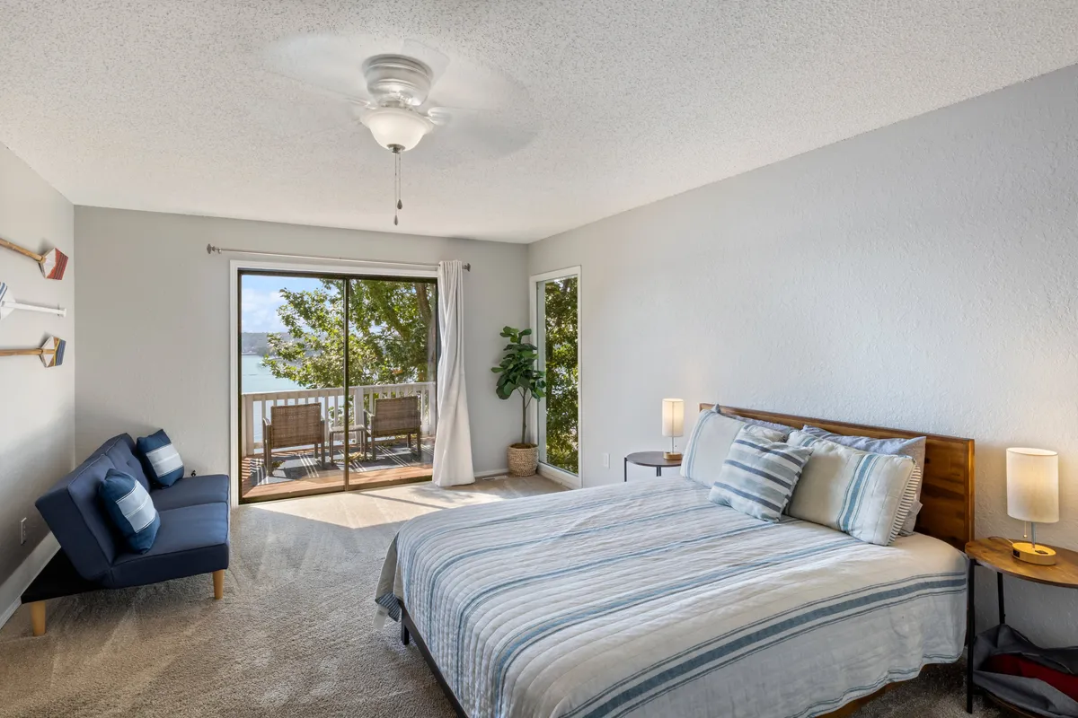 A light-filled bedroom with a comfortable bed, a sofa, a potted plant, and a nightstand. The window offers a lovely view, and the ambiance is calm and inviting—perfect for relaxation and enjoying a morning coffee.