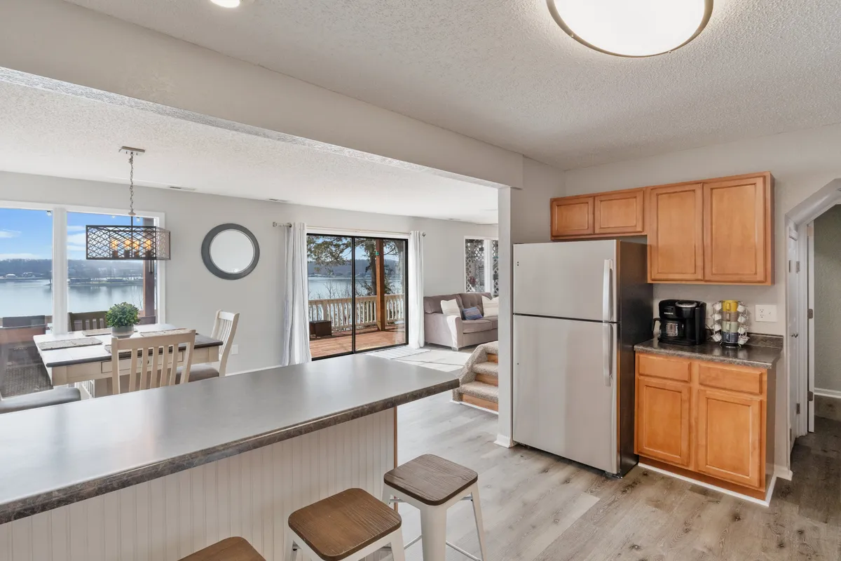 A wide-angle perspective of the main social area, including a kitchen island, dining table with chairs, and a living area with a comfortable couch. Large windows frame a stunning lake view, and the layout highlights the seamless flow between spaces.