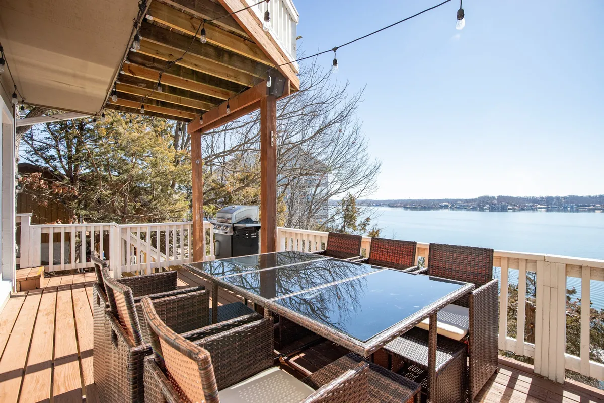 An outdoor dining table set on a deck with string lights overhead, overlooking a tranquil lake. The scene suggests intimate gatherings, sunset views, and leisurely meals under the open sky.