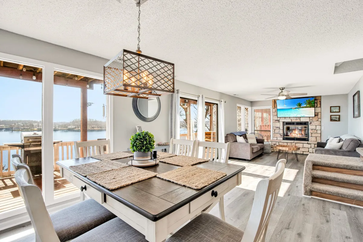 This image showcases an inviting open-plan space featuring a dining table, a cozy fireplace, and large windows offering expansive views of the lake. Comfortable seating areas are arranged for gathering and relaxation, and the entire room is bathed in natural light from the floor-to-ceiling windows.