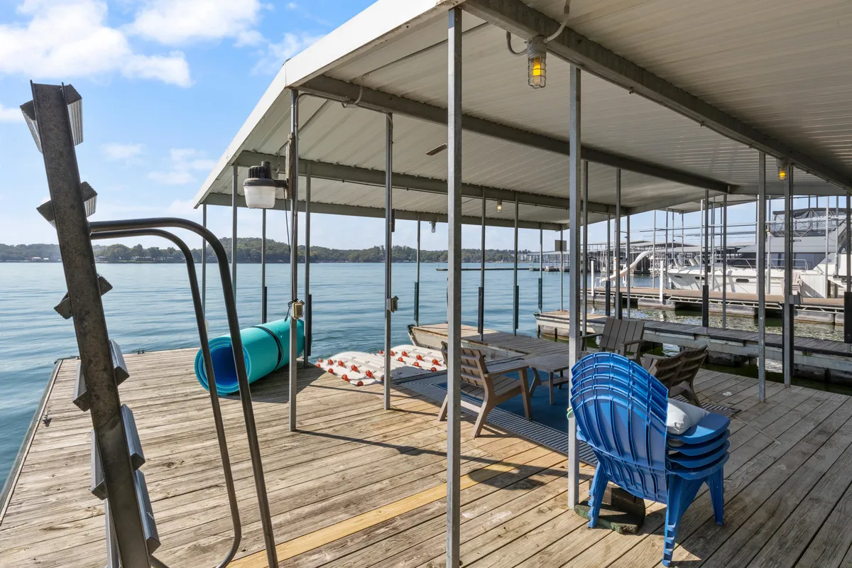 A wooden dock furnished with blue chairs and a table with cushions overlooks the calm surface of the lake. The dock is surrounded by water, creating an inviting space for relaxing, fishing, or socializing outdoors. The setting hints at proximity to attractions by water and the ability to take in scenic views day or night.