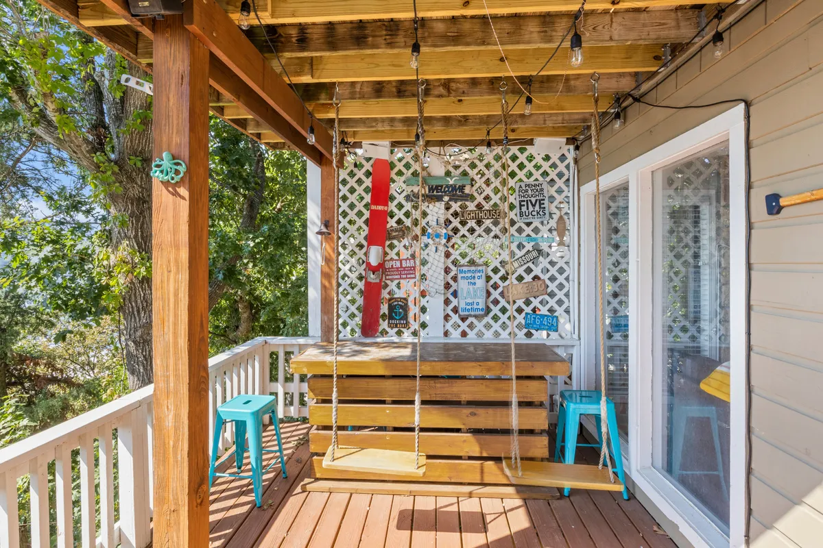 This cheerful exterior scene features a wooden swing table, blue chairs, hanging lights, and playful decorative signs—all set on a partially covered deck. The setup encourages socializing, family board games, and memorable al fresco meals. The natural surroundings and festive touches create a fun, family-friendly space for gatherings at any time of day.