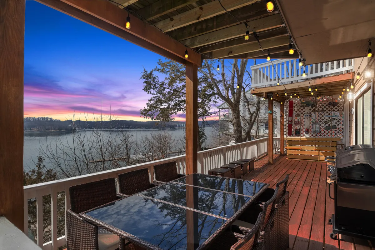 The image showcases a spacious outdoor dining area overlooking the lake, with a large table set for gatherings, warm decorative string lighting, a grill ready for barbecues, and breathtaking views of the water at sunset.