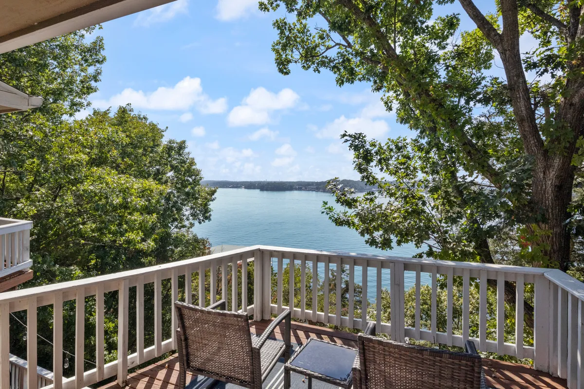 A tranquil deck seating area overlooks the lake, with comfortable outdoor chairs and a table set among surrounding trees. The vantage point highlights the peacefulness of morning coffee or evening gatherings, with the sunrise or sunset reflected over the water. The natural privacy creates an intimate, serene setting.